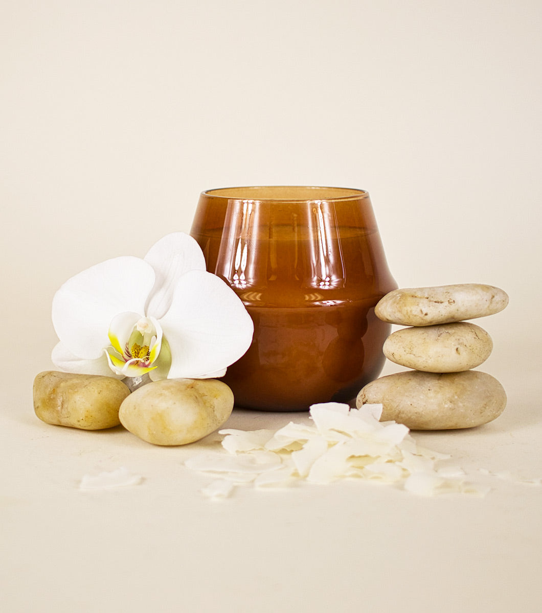 large brown amber and coconut scented candle pictured with rounded stones, a white orchid and chaved coconut